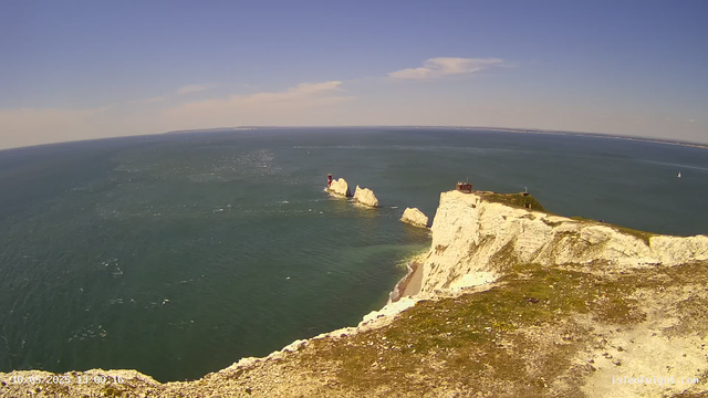 A high-angle view from a webcam shows white chalk cliffs extending into the ocean, with some small patches of grassy land on top. In the distance, there are large rock formations in the water and a lighthouse on a small headland. The sky is mostly clear with a few scattered clouds.