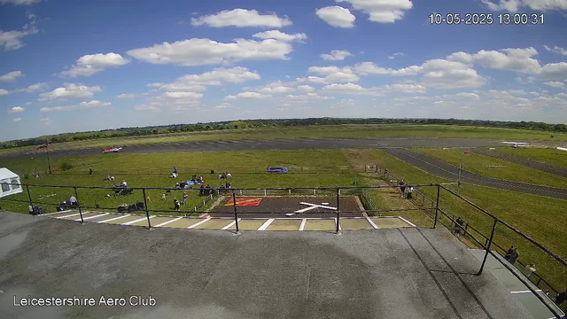 A wide view of an airfield with a grassy area and a racetrack in the background. Several people and vehicles are scattered around the grassy area, with some sitting and some standing near a fence. The weather is clear with a blue sky and scattered white clouds. The timestamp in the top right corner reads "10-05-2025 13:00:31," and the bottom left corner has the text "Leicestershire Aero Club."