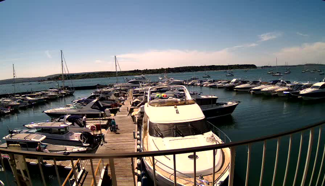 A marina filled with various boats and yachts docked along wooden piers, with more boats sailing in the distance on the water. The sky is partly cloudy, and there is a distant shoreline with some greenery across the water. The photo is taken from a vantage point behind a black metal railing.