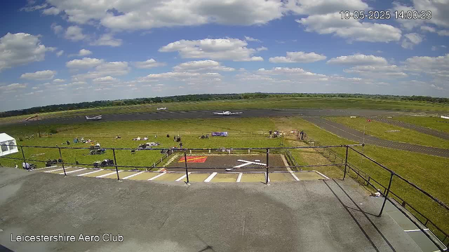 Aview from a webcam at Leicestershire Aero Club showing the runway and aircrafts parked near the edge of the airfield. The sky is partly cloudy with scattered fluffy white clouds. In the foreground, there is a platform or balcony with a railing, and below it are some parking spaces and a small fenced area. People and equipment are scattered across the grassy area near the runway. In the background, the runway extends into the distance, with a few aircraft taxiing or parked on the tarmac. The date and time are displayed in the upper right corner as 10-05-2025 14:00:23.
