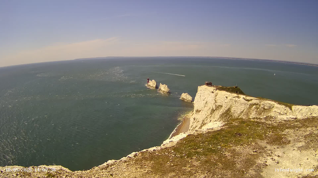 Aerial view of white chalk cliffs overlooking the sea with a grassy top, a small building near the edge, and large rock formations with a lighthouse on one of them protruding into the water. Several boats and ships are visible in the distance on the water. The sky is clear and blue.
