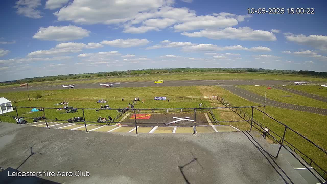A view from a webcam at Leicester Shire Aero Club showing a small airport with a grassy area in the foreground and a runway in the background. Several small aircraft are on the tarmac and taxiways, with some parked and others moving. There are people walking and sitting on benches near the grassy area. The sky is mostly sunny with scattered clouds. The timestamp in the upper right corner reads "10-05-2025 15:00:22".