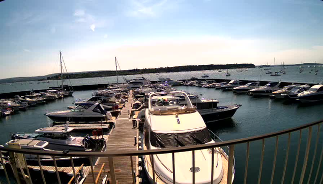 A marina with numerous docked boats and yachts, extending out into the calm water under a clear sky. The boats are lined up along wooden piers, with some boats appearing larger and more luxurious. In the background, there is a distant landmass with sparse vegetation. A curved metal railing is visible in the foreground.