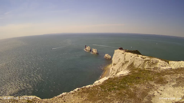 A high-angle view of white chalk cliffs extending into the sea, with a small building on top of the closest cliff, during daytime with clear skies. Several sailboats and boats are visible on the water.
