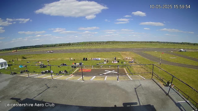 A view from a webcam at Leicester Aeroclub shows a sunny day with a clear blue sky and scattered white clouds. In the foreground, there is a paved area with a railing, some parked vehicles, and a few people sitting on the ground or standing near the railing. Beyond this, there is a grassy field with more people sitting on blankets or on the grass. Further back, a small aircraft is taking off or landing on a runway. In the distance, another aircraft is visible on the tarmac, and the horizon shows a gentle landscape with trees and low hills. The timestamp on the image indicates the date as October 5th, 2025, and the time as 15:59.