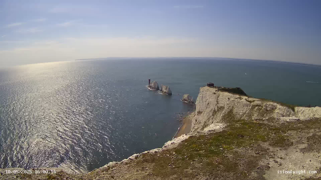Aerial view of the white chalk cliffs of Étretat along the coast, with a grassy area and a small building on top. The ocean extends into the distance, with a rock formation and a lighthouse on one of the rocks. The sky is clear and blue with a few wispy clouds.