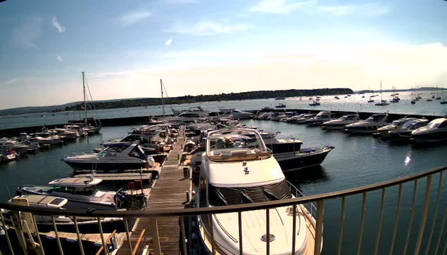 A marina filled with numerous boats and yachts docked along wooden piers, with calm water reflecting the vessels. In the background, a long breakwater extends into the water, and the sky above is partly cloudy with sunlight shining through. A curved railing is visible in the foreground.