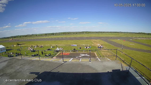 An aerial view from a webcam at LeicesterShire Aero Club shows a sunny day with a clear blue sky and a few scattered clouds. In the foreground, there is a platform or balcony with a railing, and a small parking area with a few marked spaces and a red symbol on the ground. Several people are sitting or lying on the grass, some near small tables, while a few are walking around. Beyond the grassy area, there are open fields and a track-like runway area. The scene captures a peaceful, open-air setting at the aero club on a bright day, with the date and time displayed in the top right corner as "10-05-2025 17:00:00".