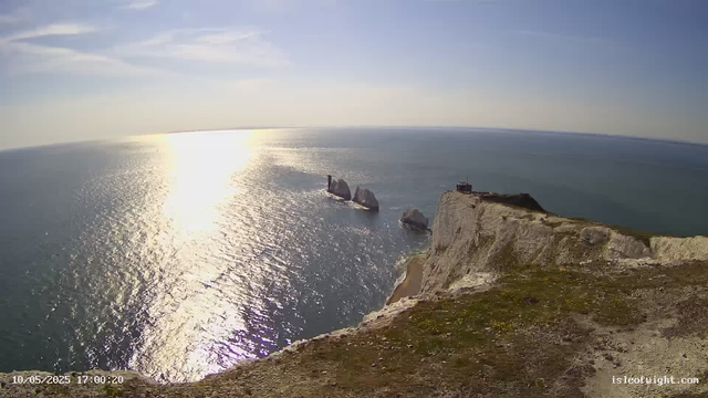 A high-angle view of white chalk cliffs overlooking the ocean at sunset. The sunlight reflects off the water, creating a shimmering effect. In the distance, there are a few sea stacks and a small building on top of the cliffs. The sky is mostly clear with some wispy clouds. The timestamp in the bottom left corner shows the date and time as May 10, 2025, at 17:00:20.