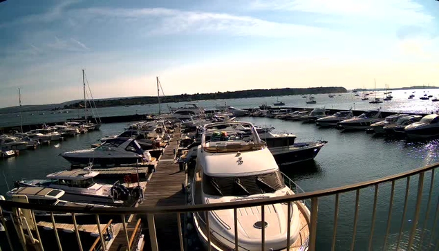 A marina filled with numerous boats moored at docks on calm water, with a clear sky and a distant landmass in the background. The photo appears to be taken from a balcony or pier with a metal railing in the foreground.