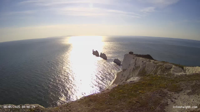 Aerial view of white chalk cliffs overlooking the ocean. The sun is shining brightly in the sky, reflecting off the water, creating a shimmering path. There are rock formations extending into the sea, with a small dark building or structure on top of the cliffs. The date and time stamp at the bottom shows "10/05/2025 18:00:29".