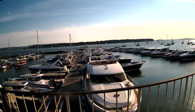 A marina filled with numerous sailboats and motorboats docked along wooden piers, with calm water reflecting the boats. In the foreground, a large white yacht is moored, and a protective metal railing surrounds the area. The sky is partly cloudy, and the distant shoreline extends along the horizon.