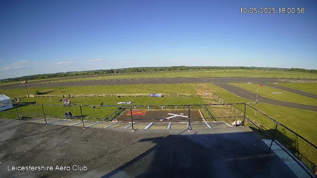 A view from a webcam of Leicestershire Aero Club shows a clear, sunny day with a bright blue sky and a few scattered clouds. In the foreground, there is a black asphalt platform with metal railings, possibly a launch or observation area. Beyond the platform, there is a grassy field with a few people and dogs, some sitting and some standing near a fenced area. The field extends to a paved runway with markings, and there are a few small structures or signs along the fencing. The landscape is flat and open, with rolling green fields and distant trees on the horizon. The date and time are displayed in the top right corner as May 10, 2025, at 6:00 PM. The bottom left corner shows the text "Leicestershire Aero Club."