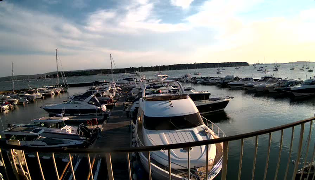 A marina with numerous boats and yachts docked at wooden piers, extending into calm water. The scene is set during the late afternoon or early evening with a partly cloudy sky and distant land visible on the horizon.