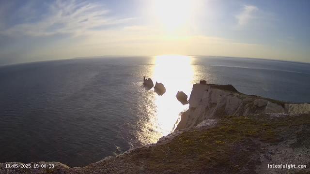 A view of a coastal landscape at sunset or sunrise, showing white chalk cliffs with a small building or structure on top. The cliffs extend into the ocean, where a few large rocks are visible near the shoreline. The water reflects the bright sunlight, creating a shimmering effect on the surface. The sky is partly cloudy with some wispy clouds. The timestamp on the image indicates the date and time as October 5, 2025, at 19:00:33.