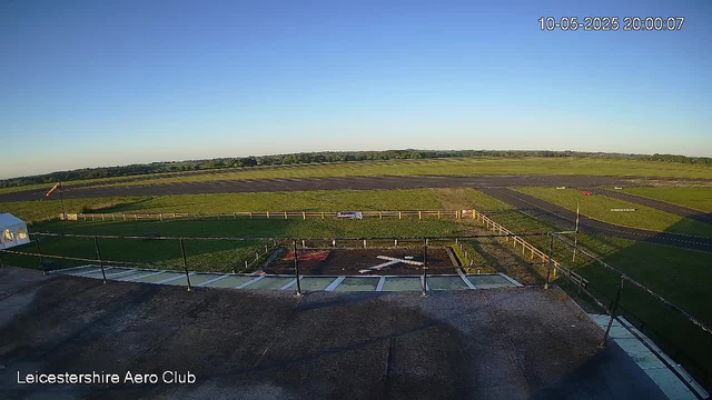 A wide view of an airfield shows a grassy runway area with some paved taxiways and an open sky. There is a small building or structure on the left with a railing in the foreground. The landscape extends into distant fields and trees on the horizon under a clear blue sky. The top right corner displays a timestamp: "10-05-2025 20:00:07," and bottom left corner reads "Leicestershire Aero Club."