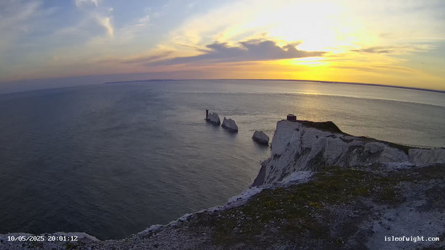 A scenic view of a coastal landscape during sunset, featuring white chalk cliffs with a building and a fence along the edge. In the sea below, there are several large white rock formations. The sky is partly cloudy with the sun near the horizon, casting a golden light over the water and cliffs.