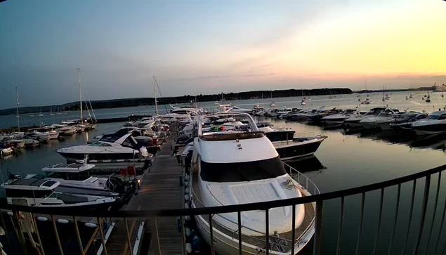 A marina at sunset with several boats docked along a wooden pier. The calm water reflects the sky's soft light, and the horizon shows a line of trees or land in the distance. The scene has a peaceful, early evening atmosphere.
