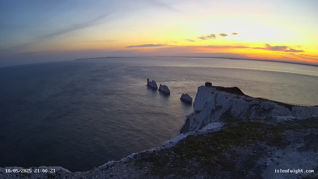 A view of white chalk cliffs overlooking the ocean at sunset, with three large sea stacks rising from the water. The sky is painted with soft hues of orange, yellow, and pink, and the horizon shows a distant landmass. The foreground features the edge of the cliffs with some grass and rocky terrain. A timestamp in the bottom left corner reads "10/05/2025 21:00:21," and a website URL "isleofwight.com" is visible in the bottom right corner.