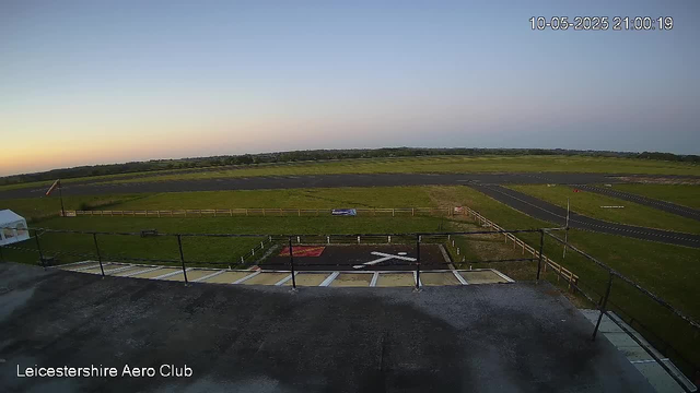 A wide view of an open airfield during sunset or sunrise, with a small helicopter landing pad marked by a white helicopter symbol in the foreground. The field is fenced and surrounded by grassy areas, with a runway extending into the distance under a clear sky. The timestamp in the upper right corner shows the date as October 5, 2025, at 9:00 PM. The text "Leicestershire Aero Club" appears in the lower left corner of the image.