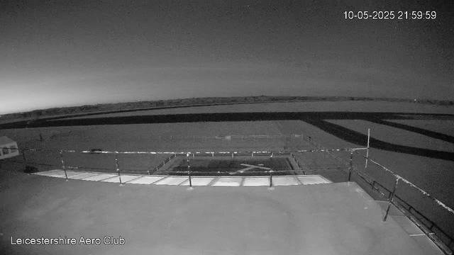 A black-and-white image from a webcam shows an outdoor area of Leicesterhire Aero Club at night, with a flat paved surface, safety railings, and a small building on the left. The sky is dark, and there are no visible people or aircraft. The timestamp in the upper right corner reads "10-05-2025 21:59:59."