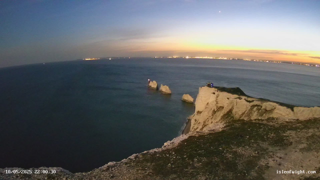 A view of white chalk cliffs extending into the ocean at sunset, with a few rocks protruding from the water near the base of the cliffs. The sky is transitioning from day to night, with a gradient of colors from light orange and yellow near the horizon to dark blue higher up. are visible. Lights from distant buildings or ships can be seen on the horizon. There is a small structure on the edge of the cliff. The bottom left corner shows a timestamp: "10/05/2025 22:00:30" and the bottom right corner has the text "isleofwight.com."