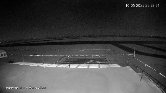 A black and white image from a webcam shows an empty airfield or tarmac area at night, with a small building or hangar visible in the far left corner. There are safety barriers and fencing surrounding what appears to be a helipad or aircraft parking area. The sky is dark with a few visible stars. The top right corner displays a timestamp of "10-05-2025 22:59:51," and the bottom left corner has the text "Leicestershire Aero Club."