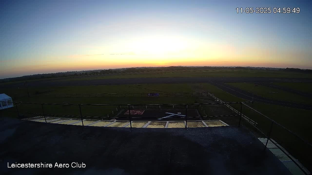A sunrise or sunset over a wide open grassy field with a track and fencing in the foreground. The sky is clear with a gradient of colors from blue to orange. The image is taken from an elevated perspective, likely a rooftop, with the text "Leicestershire Aero Club" in the bottom left corner and a timestamp in the top right corner showing the date and time as 11-05-2025 04:59:49.