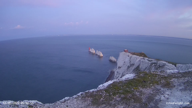 A view of white chalk cliffs on the coast extending into the ocean, with a small building and a lighthouse on the edge. Large rock formations are visible in the water near the cliffs, and the sky is partly cloudy with soft pastel colors.