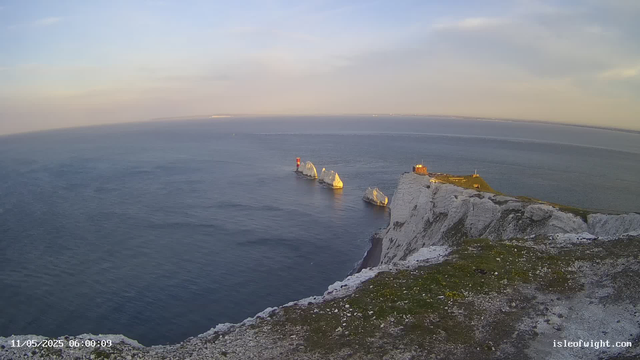A view of white chalk cliffs rising sharply from the sea, with large white rock formations in the water near the base of the cliffs. A small building and a lighthouse are situated on the edge of the cliff, overlooking the ocean. The scene is bathed in soft, golden light, suggesting early morning or late afternoon. The sky is partly cloudy with patches of blue.