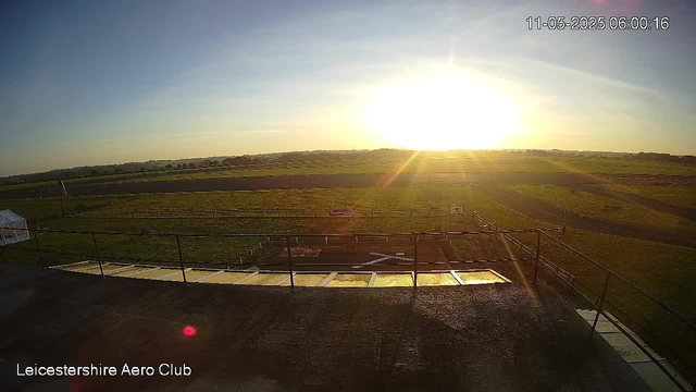 A view of a flat, grassy field during sunrise or sunset, with the sunlight creating a bright glow on the horizon. The scene includes fencing, some small structures, and a clear sky with a few faint clouds. The timestamp in the upper right corner reads "11-05-2025 06:00:16," and at the bottom left, there is text that says "Leicestershire Aero Club."