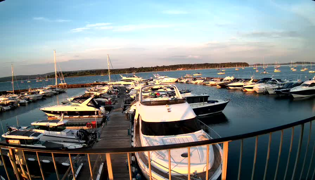 A view of a marina with numerous yachts and boats docked along wooden piers. The boats are of various sizes, with some large and others smaller. The water is calm, and the sky is partly cloudy with a hint of the setting sun, casting a warm glow over the scene. In the background, there is a tree-lined shoreline and more boats floating on the water. A metal railing curves around the foreground, indicating the photo was taken from a balcony or walkway above the marina.