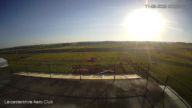 A wide view of an open, grassy airfield under bright sunlight with clear skies. In the foreground, there is a paved area with a railing and some metallic panels, possibly a viewing platform or tarmac. The background shows expansive fields, some fencing, and distant low hills on the horizon. The sun is shining brightly from the right side of the image, creating glare and long shadows. The text "Leicestershire Aero Club" is visible in the bottom left corner, and the timestamp in the upper right indicates it was taken on November 5, 2025, at 7:00 AM.