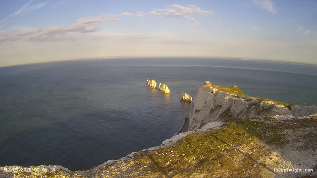 A panoramic view of white chalk cliffs with patches of green grass extends along the coastline, with the sea stretching out to the horizon under a partly cloudy sky. Large white rock formations and a lighthouse are visible near the edge of the cliffs. The scene is bathed in warm, golden sunlight.