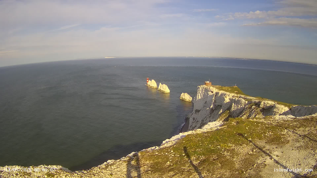 A wide view from a webcam shows white chalk cliffs with green grass at the top, jutting out into the ocean. Several white rocks and a lighthouse with red and white stripes are visible on the water near the cliffs. The sky is partly cloudy with patches of blue. The time stamp in the bottom left corner indicates the photo was taken on May 20, 2023, at 8:00 AM.