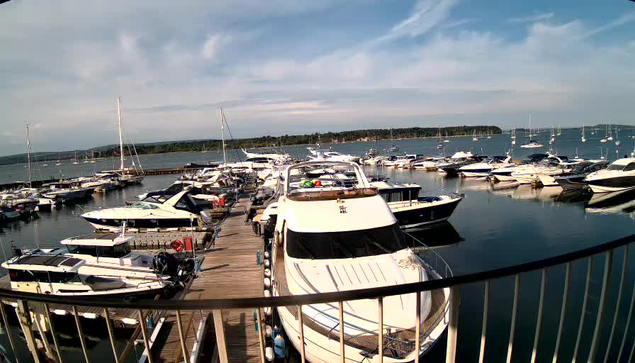 A marina with numerous boats docked along wooden piers. The boats vary in size and style, with some sailboats and motor yachts. The water is calm, reflecting the sky and boats. In the background, there is a distant shoreline with trees, and the sky above has some clouds with patches of blue.