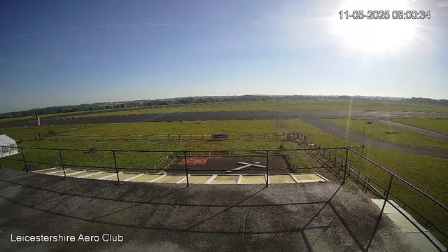 A view from a webcam shows a clear, sunny day at Leicester Shire Aero Club. In the foreground, there is a platform with black safety railings and a small helipad with a white 'H' painted on it. Beyond the platform, there is a grassy open area with a few fencing sections and a small tent on the left side. A narrow winding runway or taxiway extends across the scene, surrounded by sprawling green fields that stretch toward the horizon under a bright blue sky. The sun is high in the upper right corner, creating a glare, and a contrail from an airplane streaks diagonally across the sky. The timestamp in the top right indicates the date and time as November 5, 2025, at 8:00 a.m.