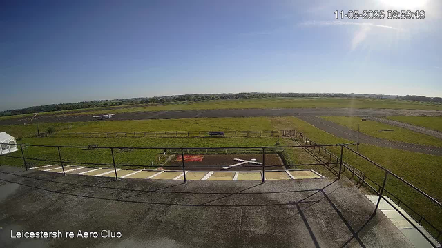 A view from a webcam showing an airfield with a clear blue sky and bright sunlight. In the foreground, there is a balcony with a railing and some parking spaces underneath. Beyond the balcony, there is a grassy area with some paved runways or taxiways. In the distance, there are small aircraft parked near the grass, and further away, a flat landscape stretches out to the horizon. The timestamp on the top right corner reads "11-05-2025 08:59:49" and the bottom left corner displays "Leicestershire Aero Club."
