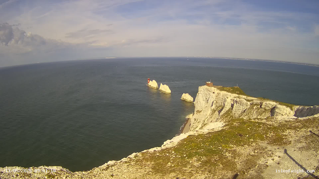 A panoramic view of a rugged coastline featuring white chalk cliffs topped with grassy patches. In the ocean below, several large chalk rock formations are visible, with one of them having a red and white lighthouse on it. The sky above is partly cloudy with blue patches, and the distant horizon stretches across the image.