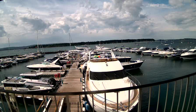A marina with numerous boats docked along a wooden pier, extending into calm water. The scene is under a partly cloudy sky, and a railing curves along the foreground. The boats vary in size and design, with some having covered cabins and others open.