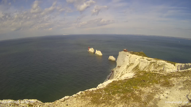 A high-angle view of white chalk cliffs overlooking the ocean, with a small lighthouse and rocky stacks in the water. The sky is partly cloudy with patches of blue.
