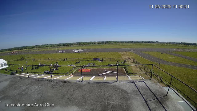 A clear day at Leicesterhire Aero Club showing a grassy area with parked motorcycles, a small tent, and a runway in the background. Two small airplanes are parked on the tarmac beyond the grassy area. The sky is blue with no clouds. The timestamp in the top right corner reads "11-05-2025 11:00:01."