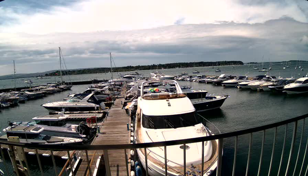 A marina with a wooden dock extending into the water, lined with various boats and yachts tethered to the sides. In the foreground, a large white yacht is prominently positioned. The background features more boats floating on calm water, a distant shoreline with some trees, and a cloudy sky overhead.