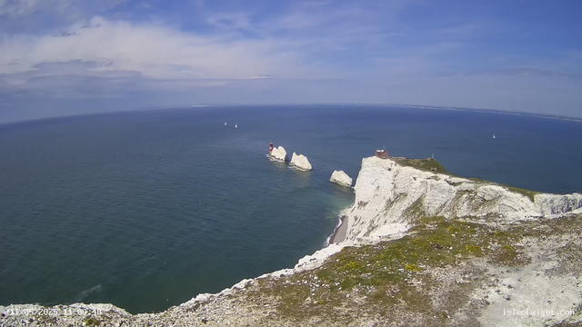 A panoramic view of white chalk cliffs along a coastline with green patches, overlooking the blue ocean under a partly cloudy sky. In the distance, there are a few sailboats and a small lighthouse on a rock formation in the sea. The cliffs have a small building and a lighthouse on top.