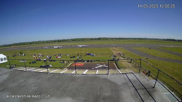 A clear day at Leicestershire Aero Club showing a grassy field with people sitting and standing, some near tents and equipment. The tarmac runway extends across the background with three small airplanes parked on it. The horizon features a line of trees under a blue sky, and there is a timestamp in the upper right corner reading "11-05-2025 12:00:25."