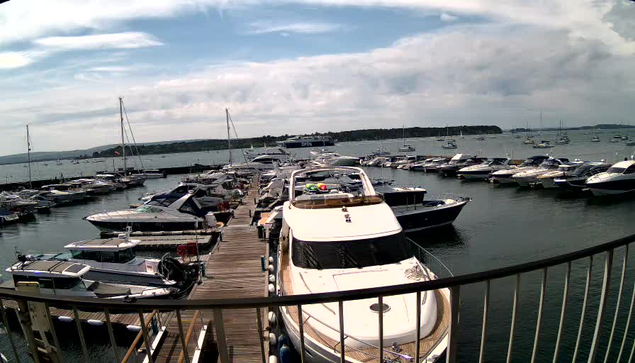 A marina filled with various boats and yachts docked along a wooden pier. The water is calm, and several sailboats are anchored further out in the bay, with a distant shoreline and some buildings visible under a partly cloudy sky.
