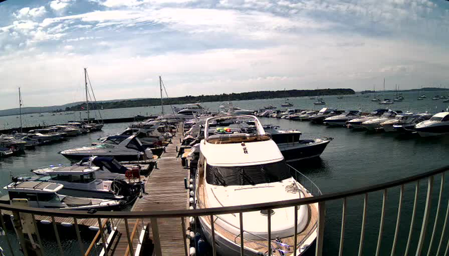 A marina filled with boats docked along wooden piers. In the foreground, a large white boat is prominently centered, with smaller boats surrounding it. The water is calm, and in the background, there is a distant shoreline with more boats scattered across the horizon. The sky is partly cloudy with patches of blue visible.