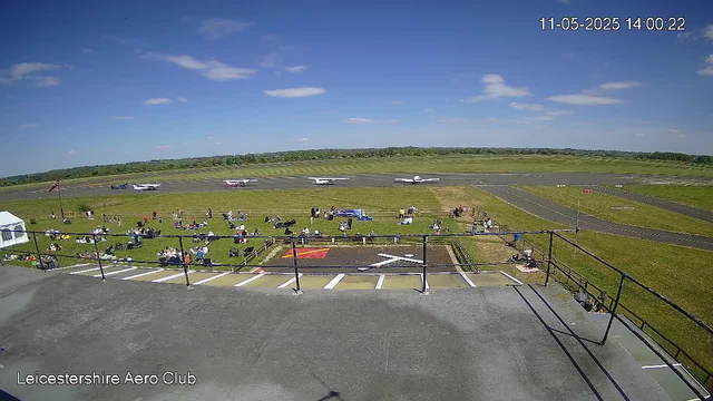 A view from a webcam at Leicestershire Aero Club shows a grassy area with people sitting and standing, some on blankets, near the runway. Four small aircraft are on the runway in the background, and the sky is partly cloudy with scattered clouds and a bright blue background. The timestamp indicates the date and time as 11-05-2025 at 14:00:22.