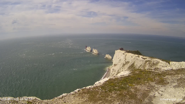 A panoramic view of white chalk cliffs rising sharply from the sea, with a small structure on top of the nearest cliff. Several large white rock formations jut out from the water further out to sea. The sky is partly cloudy with patches of blue, and the ocean has gentle waves. A boat speeds across the water, leaving a white trail behind it. The timestamp shows the date and time: 11/05/2025, 14:00:51.