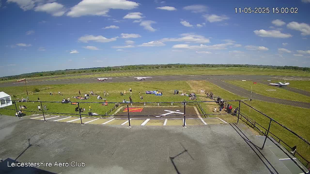 A view of a small airfield on a bright, sunny day with a partly cloudy sky. In the foreground, there is a viewing platform with a black railing, and some parked motorcycles and bicycles beside it. People are walking and sitting on the grass, some near tents and benches. In the middle distance, there are two planes on the runway, one facing left and the other facing right, with a few more planes in the background on the tarmac. The landscape is flat with green fields stretching to the horizon. The image has a timestamp in the top right corner and the text "Leicestershire Aero Club" at the bottom left.